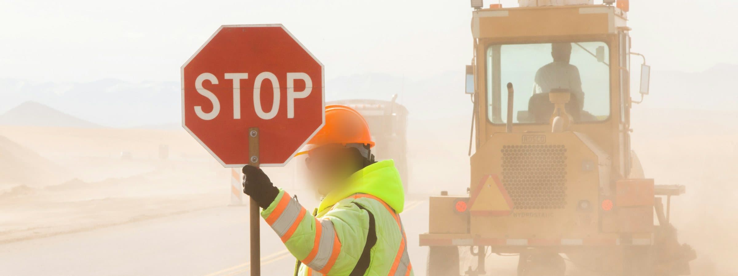 A traffic control person in a high-visibility jacket holding a stop paddle on a dusty highway, a grader working behind them.