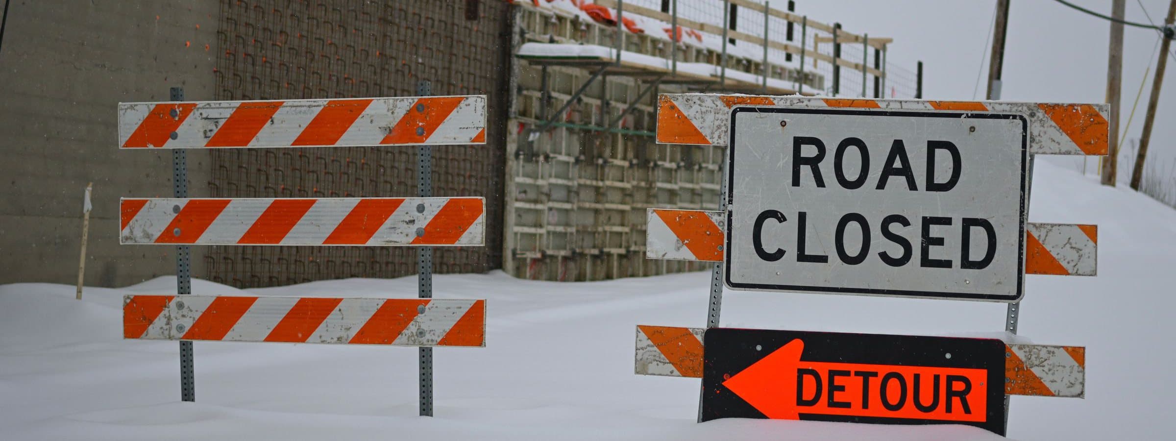 Road closed and detour signs on striped barricades, half-buried in snow at a winter work site.
