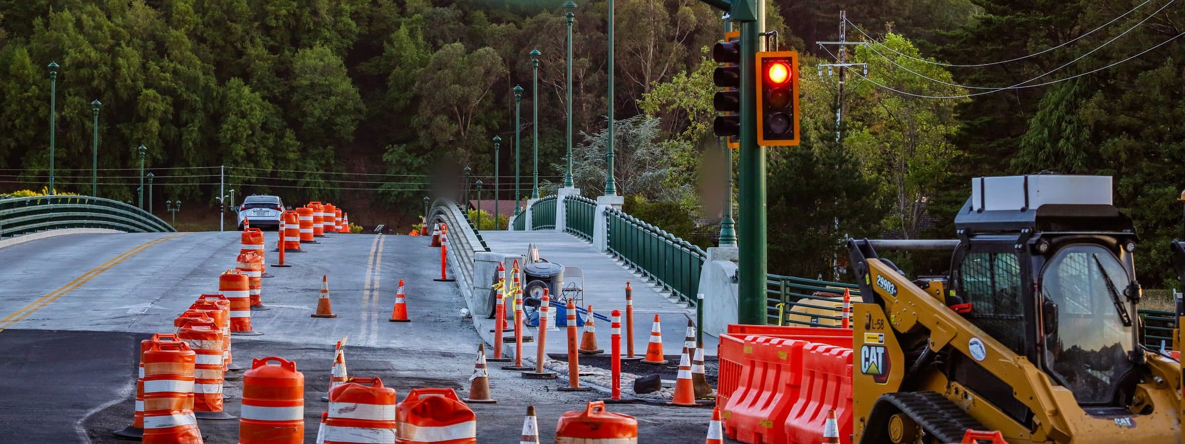 Orange barrels and cones tapering a closed lane across a bridge, temporary signals showing red, forested hillside beyond.