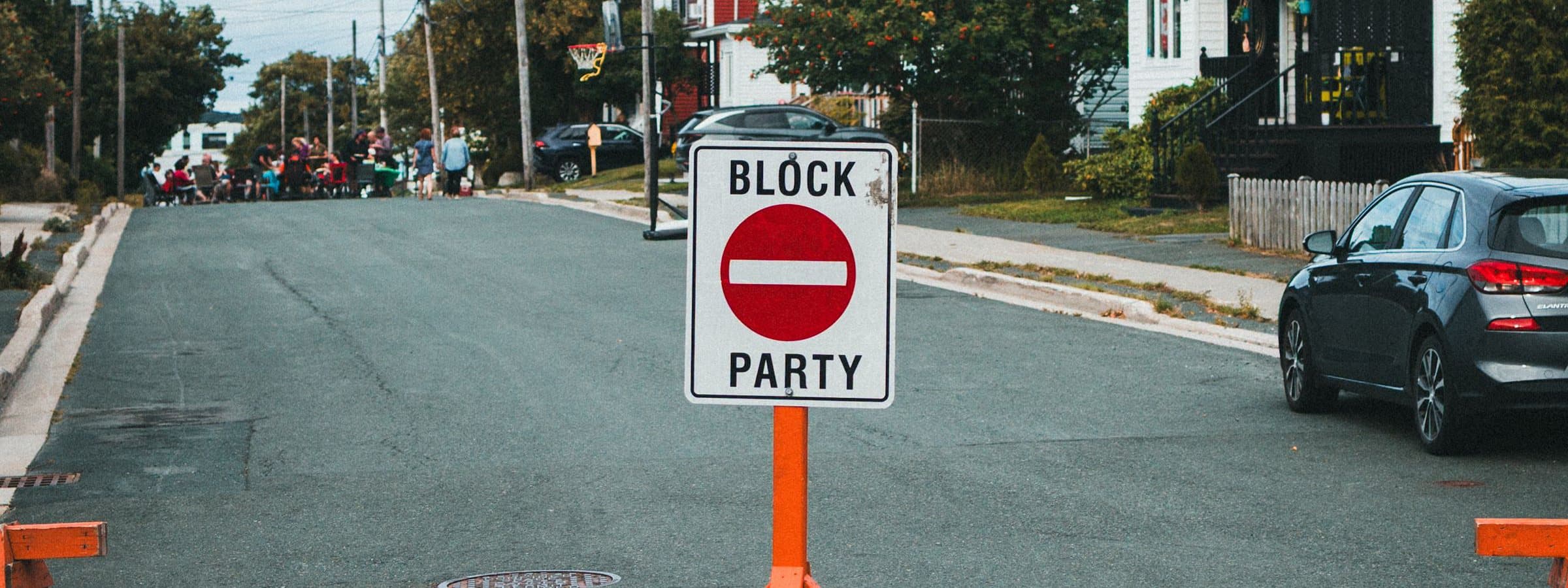 A residential street closed for an event, orange trestle barricades either side of a no-entry sign, a crowd gathered up the road.