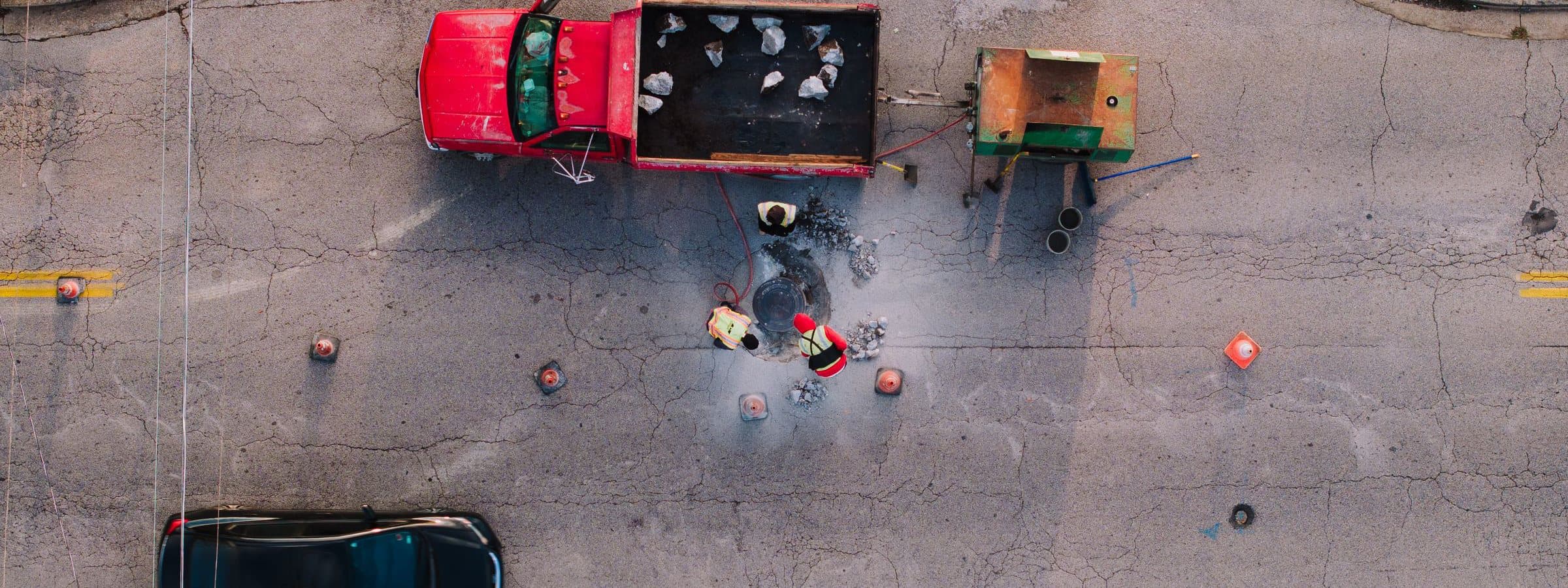 Overhead view of a road repair: a truck, a tar kettle and cones laid out around an excavation across one lane.